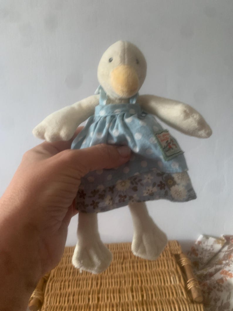 Pu&ograve; includere: Una bambola di peluche a forma di oca con corpo color crema, becco giallo e vestito blu a pois bianchi. L'abito presenta un motivo floreale sul fondo. La bambola ha le ali spiegate ed &egrave; alta circa 20 cm.
