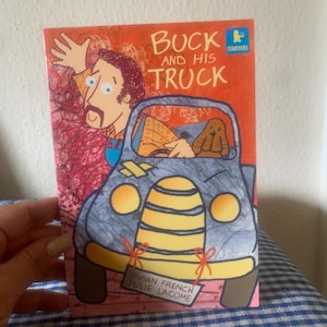 Könnte beinhalten: Ein Kinderbuchcover mit dem Titel "Buck and His Truck" zeigt eine Illustration eines Mannes, der aus einem blauen Lastwagen winkt. Das Buch hat einen orangefarbenen Hintergrund mit dem Wort "Starters" in einer blauen Box. Die Namen der Autoren, Vivian French und Julie Lacome, sind ebenfalls sichtbar.