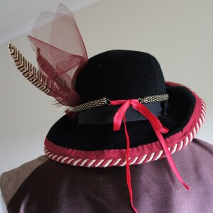 May include: A black felt hat with a wide brim, trimmed with a red and white rope. The hat has a red ribbon tied at the back and a feather and a piece of red netting on the side.