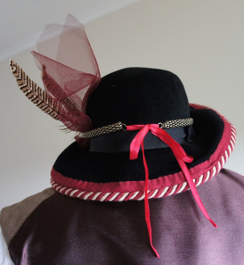 May include: A black felt hat with a wide brim, trimmed with a red and white rope. The hat has a red ribbon tied at the back and a feather and a piece of red netting on the side.