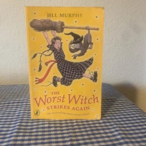 May include: A copy of "The Worst Witch Strikes Again" by Jill Murphy. The cover is yellow with an illustration of a young witch on a broom, and a black cat. The book is a children's classic.
