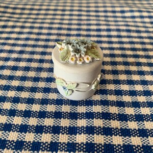 Può includere: Un piccolo cofanetto in ceramica bianca con un design floreale. Il coperchio è decorato con fiori blu e gialli e foglie verdi.