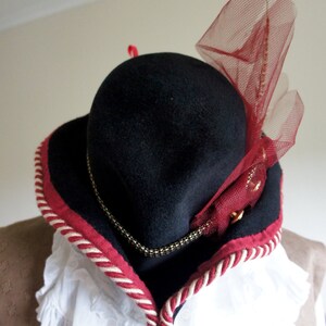 May include: A black felt hat with a red and white trim and a red tulle bow. The hat has a wide brim and a tall crown.