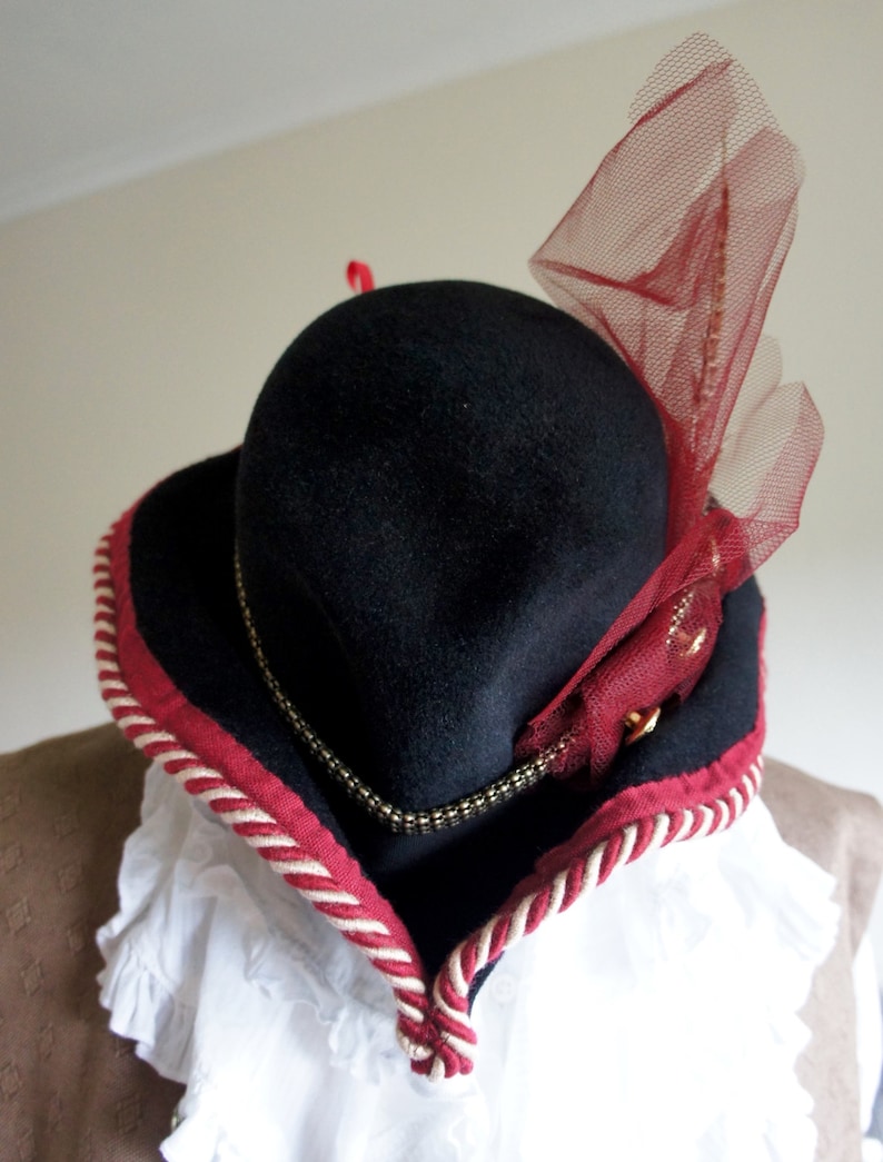 May include: A black felt hat with a red and white trim and a red tulle bow. The hat has a wide brim and a tall crown.