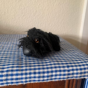 May include: Black plush toy dog with brown eyes. The dog is lying on a blue and white checkered fabric.
