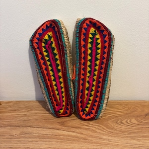 May include: A pair of handmade slippers with detailed embroidery. The slippers display a vibrant mix of colours: red, orange, yellow, blue, and pink. The design incorporates geometric patterns and a straw-like border.