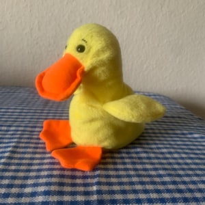 May include: A plush yellow duck toy with an orange beak and feet. The duck has small black eyes and a soft, textured body. It is sitting on a blue and white checkered surface.