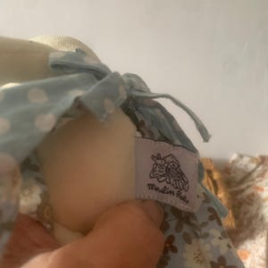 Pu&ograve; includere: Una bambola di peluche morbida con un vestito azzurro a pois bianchi e motivi floreali. La bambola ha un'etichetta con la scritta "Moulin Roty". La testa della bambola &egrave; color crema.