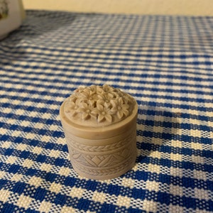 Könnte beinhalten: Eine kleine, runde, beige Schmuckdose mit einem Blumendesign auf dem Deckel und einem geschnitzten Muster um die Basis. Die Dose steht auf einer blau-weiß karierten Tischdecke.