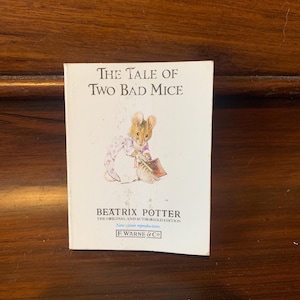 May include: A vintage book cover featuring a white background with the title "The Tale of Two Bad Mice" in black text. The cover also includes an illustration of a mouse wearing a pink dress and holding a piece of cake. The author, Beatrix Potter, is listed below the title, along with the text "The Original and Authorized Edition" and "New colour reproductions" in smaller font. The publisher, F. Warne & Co, is listed at the bottom.