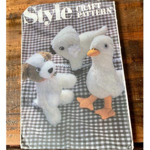 May include: A vintage craft pattern book titled "Style Craft Pattern" featuring patterns for stuffed animal toys. The toys include a dog, a duck, and a swan, all made of white fabric with colorful accents. The background is a checkered pattern.