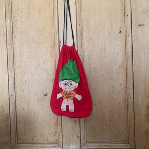 Op de afbeelding: Rode trekkoordtas met een groene, harige troll-cartoonfiguur die een oranje shirt draagt met de tekst "Veel geluk".