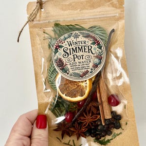 Pu&ograve; includere: Un sacchetto di plastica trasparente pieno di ingredienti per un simmer pot invernale. Il sacchetto ha un esterno di carta marrone con un'etichetta che recita "Winter Simmer Pot" e istruzioni per aggiungere acqua e far sobbollire a fuoco lento. Gli ingredienti includono fette d'arancia, bastoncini di cannella, anice stellato, bacche di ginepro e rametti di sempreverdi.