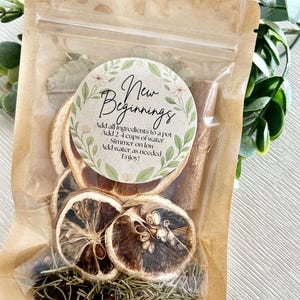 Puede incluir: Una bolsa transparente llena de hierbas secas, especias y rodajas de cítricos. La bolsa está sellada con una parte superior de papel marrón y una etiqueta circular con la inscripción "New Beginnings" e instrucciones.