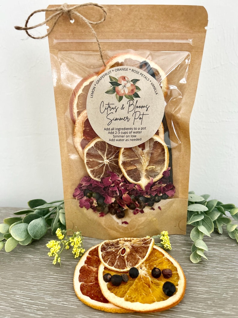 Pu&ograve; includere: Un sacchetto di carta marrone con una finestra di plastica trasparente, contenente fette di agrumi essiccati, petali di rosa e grani di pepe nero. Il sacchetto ha un'etichetta che recita "Citrus & Blooms Simmer Pot" e istruzioni per l'uso.