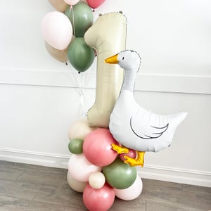 May include: A festive balloon arrangement featuring a large number one balloon in a neutral tone, a white goose balloon with yellow feet and beak, and a cluster of pink, green, and neutral-toned balloons. Perfect for a first birthday celebration.