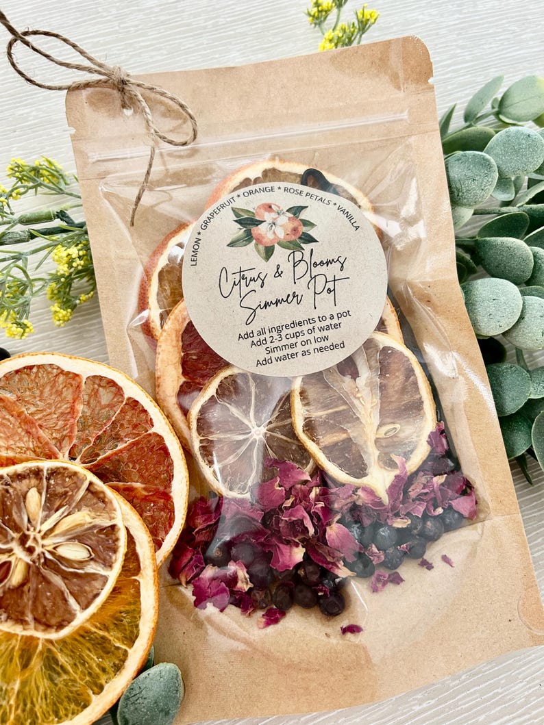 Pu&ograve; includere: Un sacchetto di carta marrone con una finestra di plastica trasparente che contiene fette di agrumi essiccati, petali di rosa e bacche nere. Il sacchetto ha un'etichetta bianca con la scritta "Citrus & Blooms Simmer Pot" e istruzioni per l'uso.