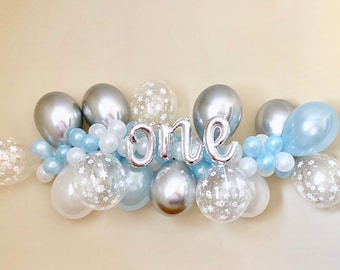 May include: A silver and blue balloon garland with the word "one" spelled out in silver balloons. The garland is decorated with snowflake patterned balloons.