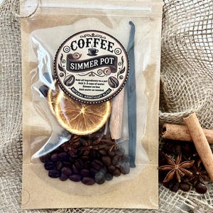 May include: A clear plastic pouch with a brown paper backing containing a coffee simmer pot blend. The label reads COFFEE SIMMER POT, NATURAL AROATHEAPY BLEND, and instructions. Inside are coffee beans, a dried orange slice, a cinnamon stick, and a vanilla bean.