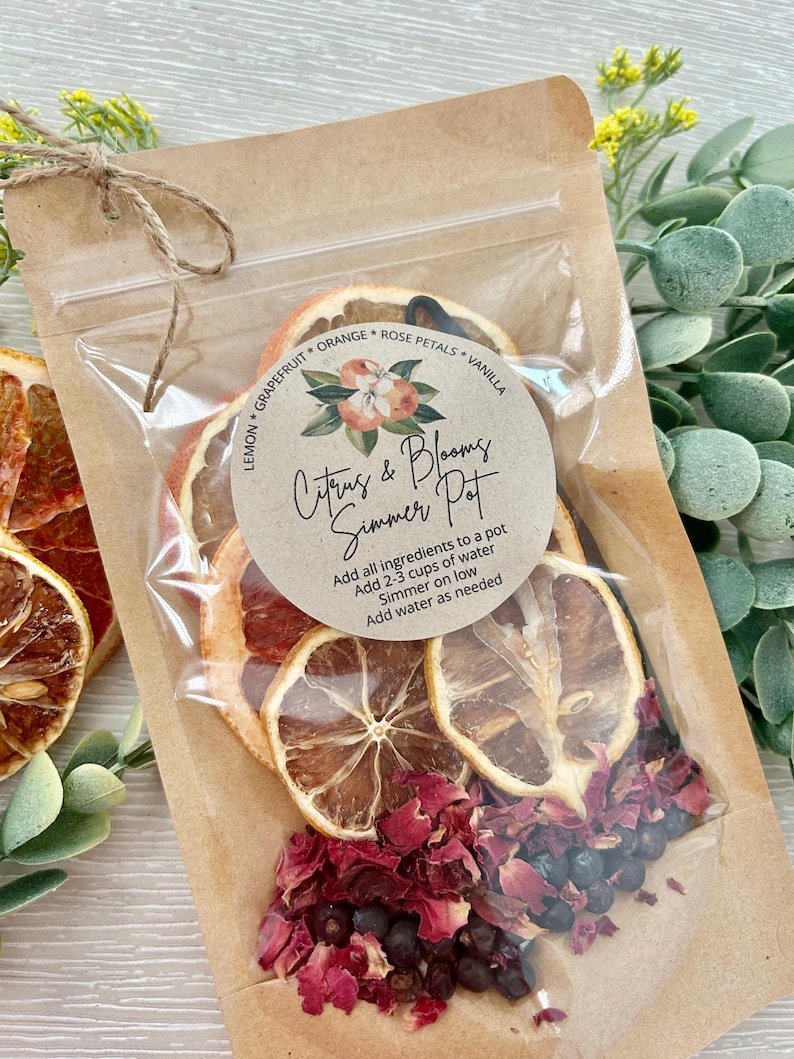 Pu&ograve; includere: Un sacchetto di carta marrone con una busta di plastica trasparente che contiene frutta di agrumi secca, petali di rosa e bacche di ginepro. Il sacchetto ha un'etichetta che recita "Citrus & Blooms Simmer Pot" e include istruzioni per l'uso.