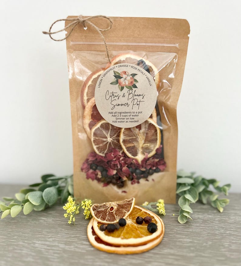 Pu&ograve; includere: Un sacchetto di carta marrone con una finestra di plastica trasparente che contiene frutta di agrumi secca, petali di rosa e bacche. Il sacchetto ha un'etichetta che recita "Citrus & Blooms Summer Pot" e include istruzioni per preparare una bevanda infusa con agrumi e fiori.