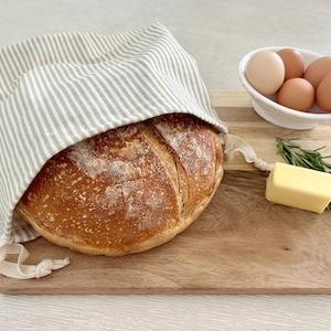 May include: A loaf of freshly baked bread is partially covered by a striped linen bread bag. The bread sits on a wooden cutting board, with a small bowl of eggs, a sprig of rosemary, and a pat of butter.