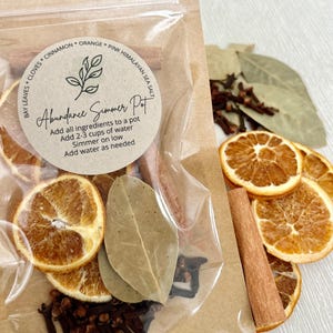 May include: A clear bag filled with dried orange slices, bay leaves, cloves, and cinnamon sticks. A round label reads "Abundance Simmer Pot" with instructions. The ingredients are for a simmer pot, a home fragrance method.