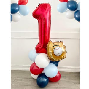 May include: A red number one balloon with a gold baseball glove and ball attached. The balloon is surrounded by blue, red, and white balloons.