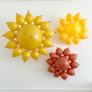 May include: Three balloon flower decorations in yellow and orange hues against a white wall. The largest is yellow, surrounded by yellow teardrop-shaped balloons. The other two are smaller, one yellow and one orange.
