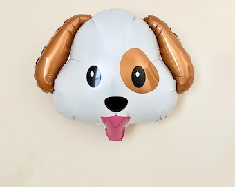 May include: A foil balloon shaped like a dog head with white fur, a brown patch over the left eye, two long brown ears, black oval eyes, a black nose, and a pink tongue sticking out from the mouth, creating a playful and expressive canine design.