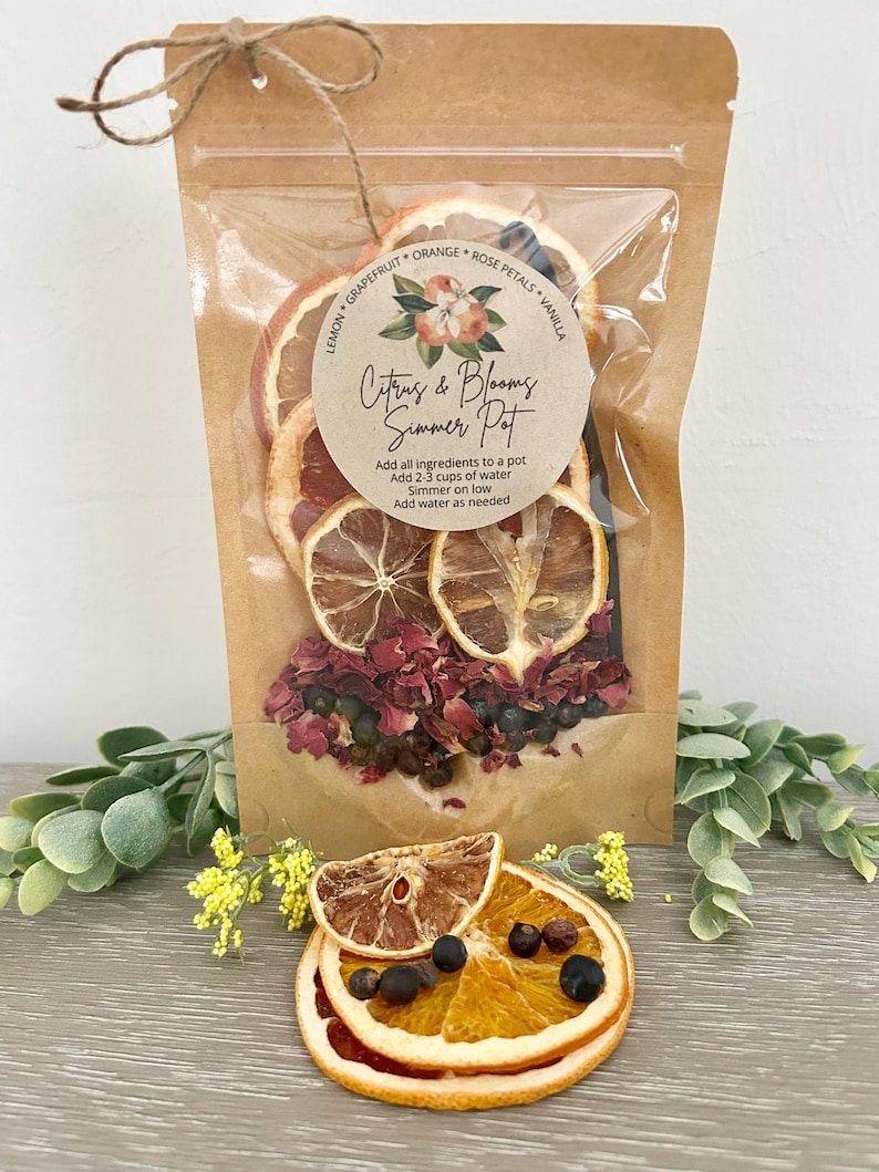 Pu&ograve; includere: Un sacchetto di carta marrone con una finestra di plastica trasparente, contenente fette di agrumi essiccati, petali di rosa, baccelli di vaniglia e bacche di ginepro. Il sacchetto ha un'etichetta che recita "Citrus & Blooms Simmer Pot" e istruzioni per l'uso.