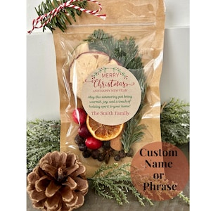 May include: A clear plastic bag filled with dried fruit, spices, and evergreen sprigs. A round label with the text "Merry Christmas and Happy New Year" is attached to the bag. The label also includes the text "May this simmering pot bring warmth, joy, and a touch of holiday spirit to your home! The Smith Family". A second round label is visible with the text "Custom Name or Phrase".