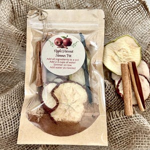 May include: A brown paper bag with a clear plastic window, containing dried apple slices, cinnamon sticks, and vanilla beans. The bag has a white label with the text "Apple Harvest Simmer Pot" and instructions for use.