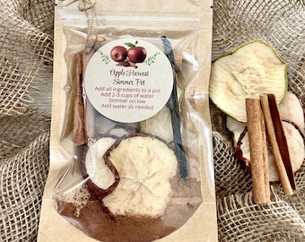 May include: A brown paper bag with a clear plastic window, containing dried apple slices, cinnamon sticks, and vanilla beans. The bag has a white label with the text "Apple Harvest Simmer Pot" and instructions for use.