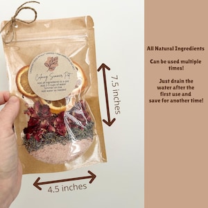 Op de afbeelding: Een bruine kraftpapieren zak met gedroogde sinaasappelschijfjes, rozenblaadjes, lavendel en roze Himalayazout. Het etiket vermeldt Calming Simmer Pot. De zak is 19,05 cm hoog en 11,43 cm breed, met tekst over de natuurlijke ingredi&euml;nten.