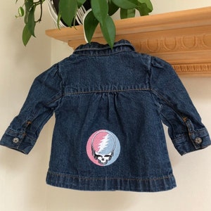 May include: A dark blue denim jacket with a button-down front and long sleeves. The back of the jacket features a round patch with a skull and lightning bolt design in pink, white, and blue. The jacket is hanging on a white hanger.
