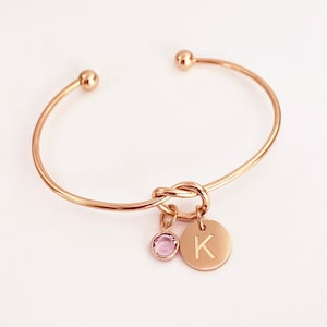 May include: A gold-colored open cuff bracelet with a knot design. It features a small, round, pink gemstone charm and a circular charm engraved with the letter "K". The bracelet has ball-shaped ends.