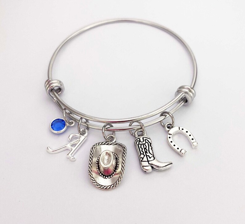 May include: A silver bangle bracelet with charms including a horseshoe, cowboy boot, cowboy hat, and the letter A. A blue gemstone is also attached to the bracelet.