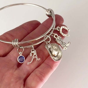 May include: Silver charm bracelet with a horseshoe, cowboy boot, cowboy hat, letter A charm, and a blue gemstone charm.