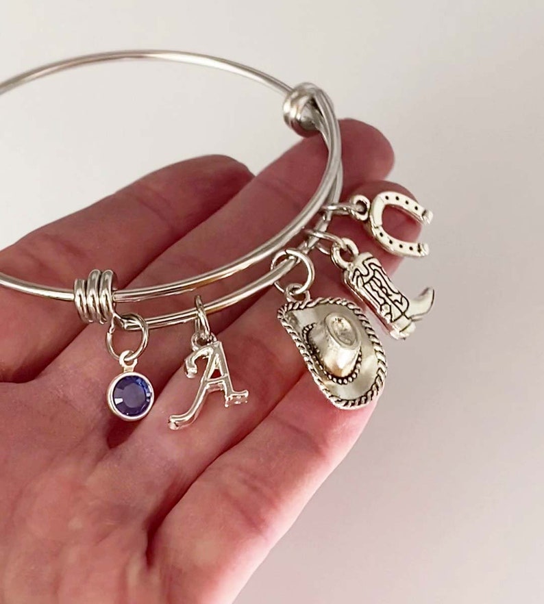May include: Silver charm bracelet with a horseshoe, cowboy boot, cowboy hat, letter A charm, and a blue gemstone charm.