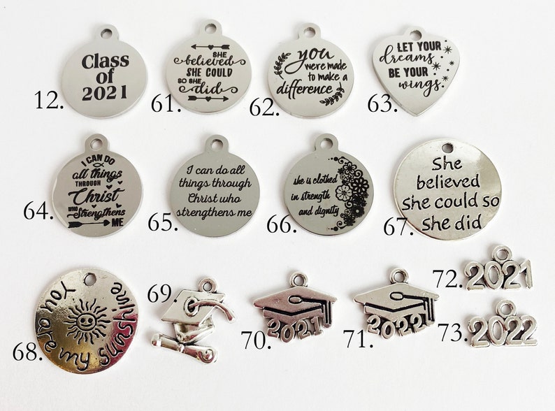May include: Assortment of silver charm pendants with inspirational quotes and graduation themes.  The charms feature phrases like "She believed she could so she did", "You were made to make a difference", and "Let your dreams be your wings".  There are also graduation caps with the year 2021 and 2022.