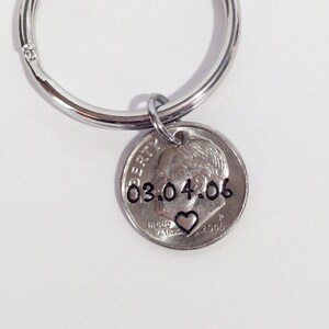 May include: A silver-colored keychain with a dime pendant. The dime is stamped with the date "03.04.06" and a small heart symbol. The keychain has a circular ring for attaching keys.
