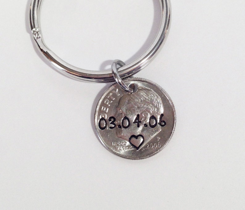 May include: A silver-colored keychain with a dime pendant. The dime is stamped with the date "03.04.06" and a small heart symbol. The keychain has a circular ring for attaching keys.