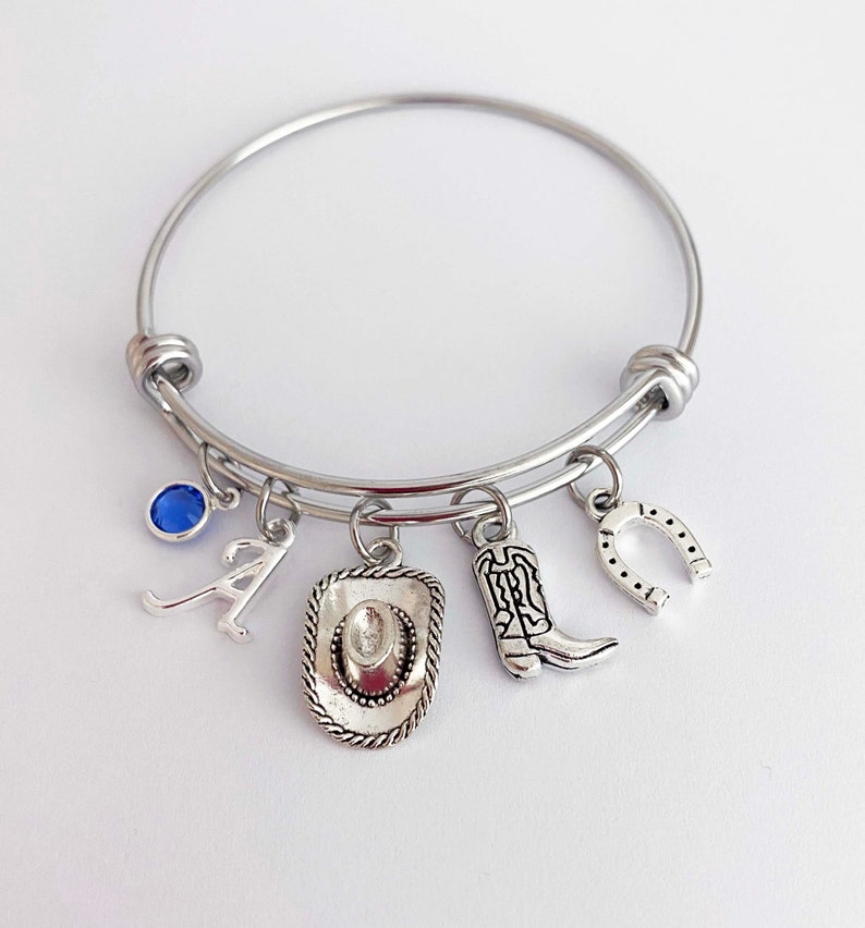 May include: A silver charm bracelet with a blue gemstone, a letter A charm, a cowboy hat charm, a cowboy boot charm, and a horseshoe charm.