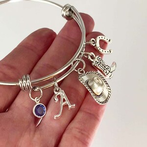 May include: Silver charm bracelet with a horseshoe, cowboy boot, cowboy hat, letter A charm, and a blue gemstone charm.