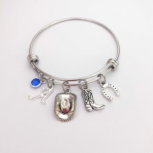 May include: A silver charm bracelet with a blue gemstone, a letter A charm, a cowboy hat charm, a cowboy boot charm, and a horseshoe charm.