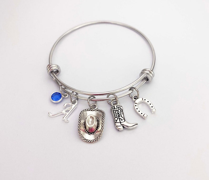 May include: A silver charm bracelet with a blue gemstone, a letter A charm, a cowboy hat charm, a cowboy boot charm, and a horseshoe charm.