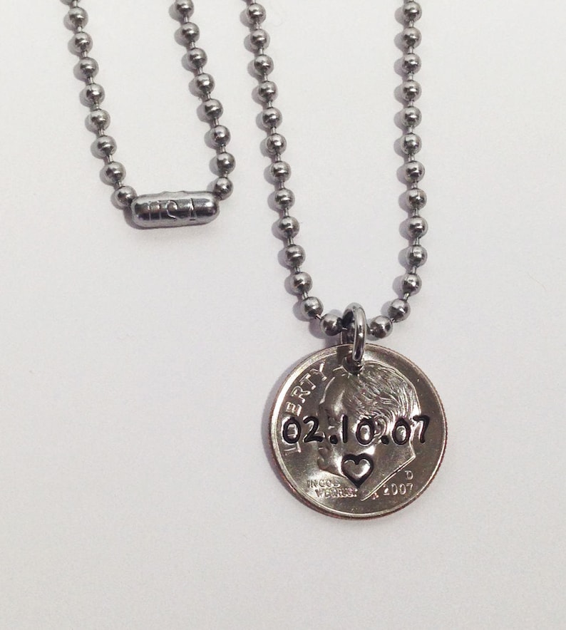 May include: A silver-colored necklace with a ball chain and a coin pendant. The coin is engraved with the date 02.10.07 and a heart symbol. The necklace is displayed against a white background.