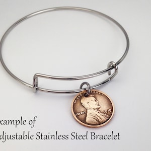 May include: A silver adjustable bangle bracelet with a copper-colored 1919 penny charm. The penny features a profile of Abraham Lincoln and the words "In God We Trust" and "Liberty".