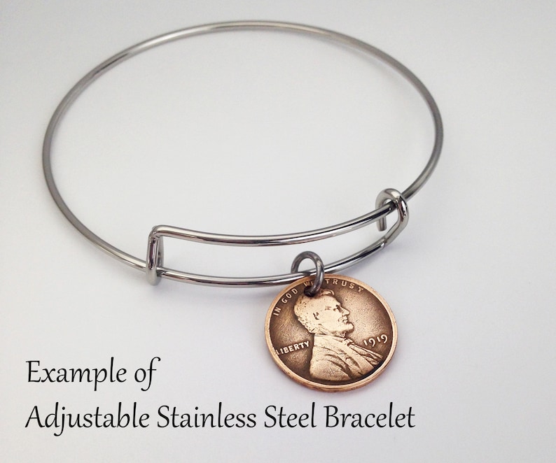May include: A silver adjustable bangle bracelet with a copper-colored 1919 penny charm. The penny features a profile of Abraham Lincoln and the words "In God We Trust" and "Liberty".